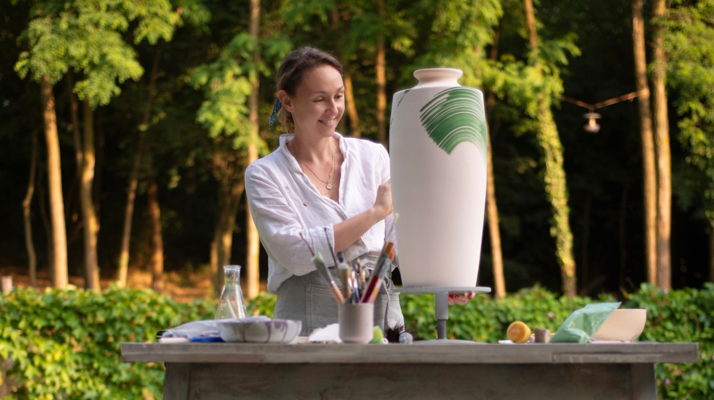 Woman working with a large ceramic vase outdoors in a forest setting